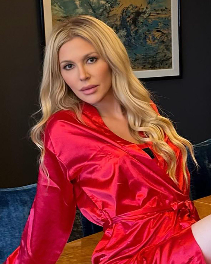 Brandi Glanville wearing a red robe, posing indoors with long blonde hair and soft makeup in front of a framed painting.