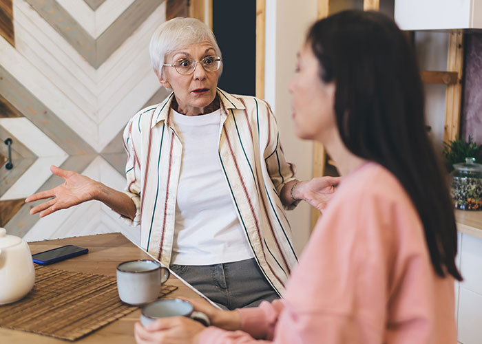 Older woman looking surprised while talking to younger woman at a kitchen table, capturing mother humiliated wedding moment.
