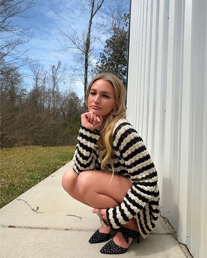 Young woman squatting outdoors wearing a striped sweater and heels, reflecting Britney Spears net worth in 2025 context.
