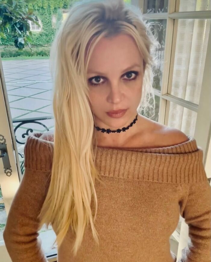 Blonde woman indoors with long hair and smoky eye makeup, wearing a brown off-shoulder top and a black choker necklace.