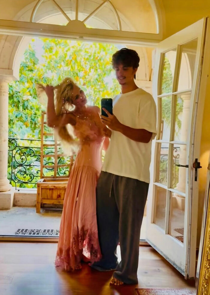 Britney Spears in a pink dress dancing playfully while a man takes a mirror selfie inside a bright room with an open door.