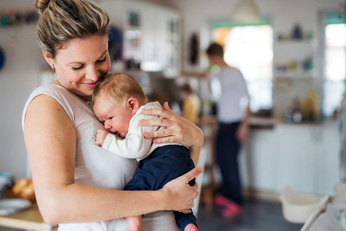 Postpartum woman holding a baby in the kitchen, showing the challenges of helping with triplets and family tension. Postpartum woman holding a baby in the kitchen, showing the challenges of helping with triplets and family tension.