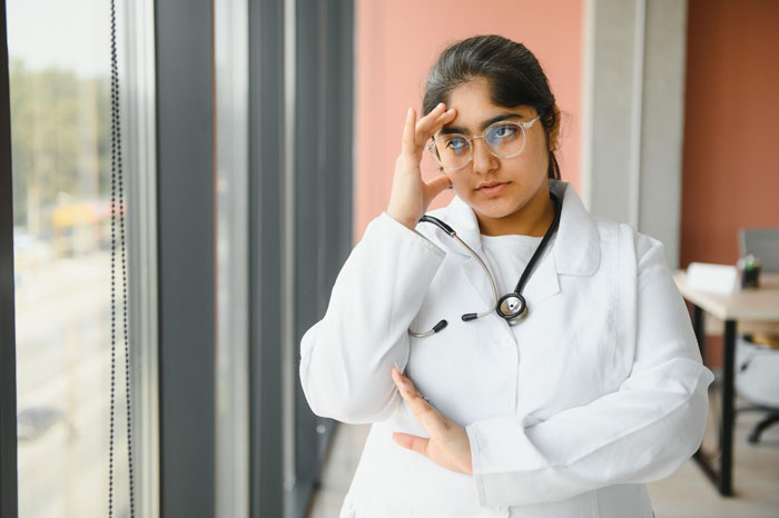 Young female doctor in white coat with stethoscope looking concerned by the window in a medical office setting Young female doctor in white coat with stethoscope looking concerned by the window in a medical office setting