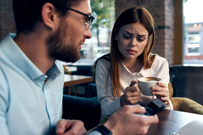 Couple in a cafe with the woman looking flabbergasted while holding a coffee cup and the man focused on his phone.
