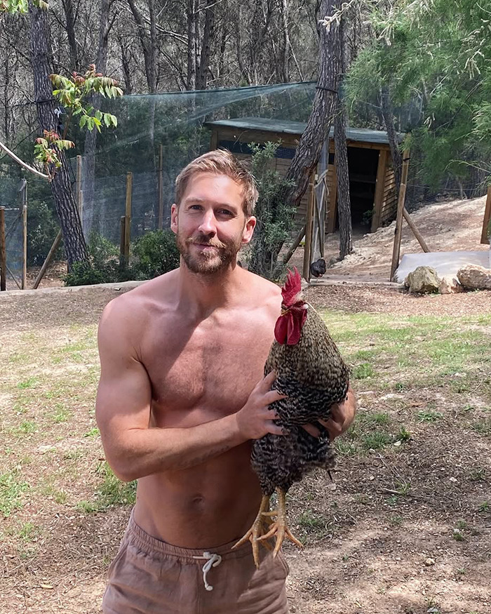 Shirtless man holding a rooster outdoors near a fenced area with trees and a wooden coop behind. - 12
