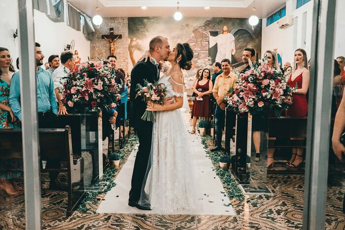 Bride and groom kissing in a decorated church aisle during wedding ceremony with guests watching and floral arrangements nearby
