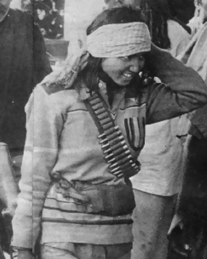Woman wearing bandolier and blindfold, smiling while standing among a group, illustrating chaotic good morally grey acts.