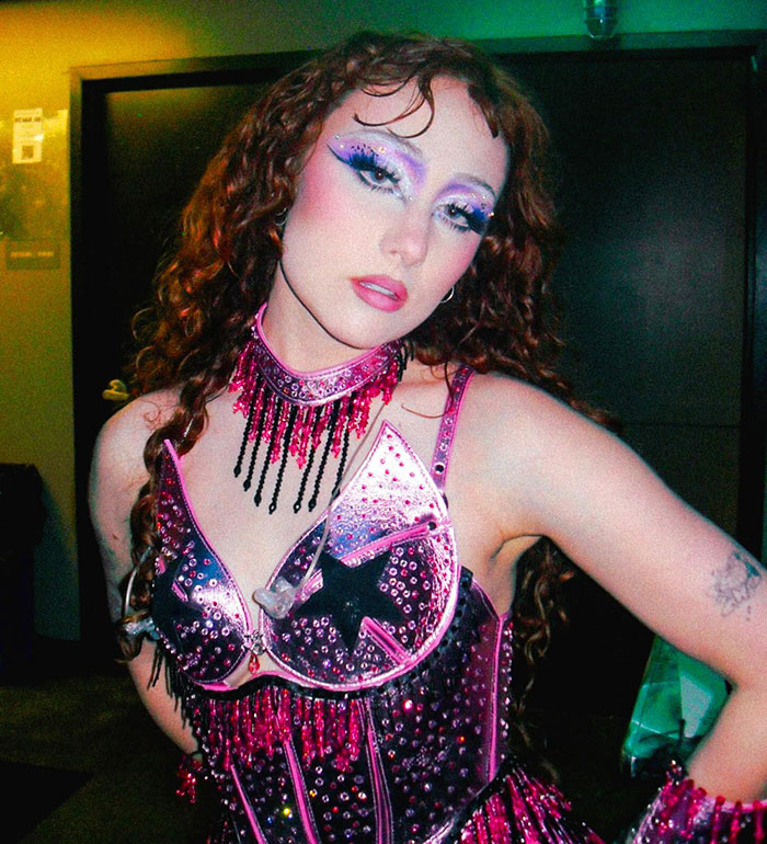 Woman with curly hair in flashy purple and pink outfit and dramatic makeup, relating to Taylor Swift cover art controversy. Woman with curly hair in flashy purple and pink outfit and dramatic makeup, relating to Taylor Swift cover art controversy.