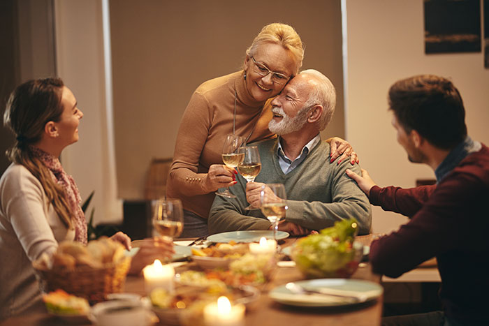 Family dinner scene with parents and a couple sharing drinks, hinting at a compliment causing tension between them.