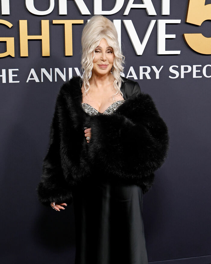Cher at an event wearing a black fur coat and glittering top, sparking concern over her plastic surgery indulgence.
