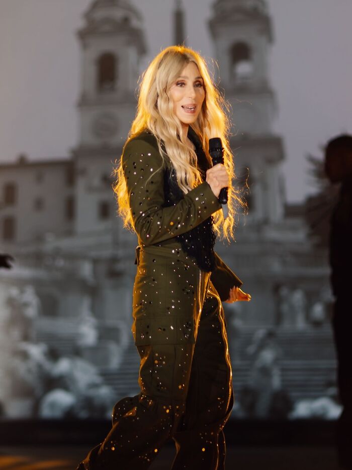Cher performing on stage with long blonde hair, wearing a studded outfit amid concerns about her plastic surgery indulgence.