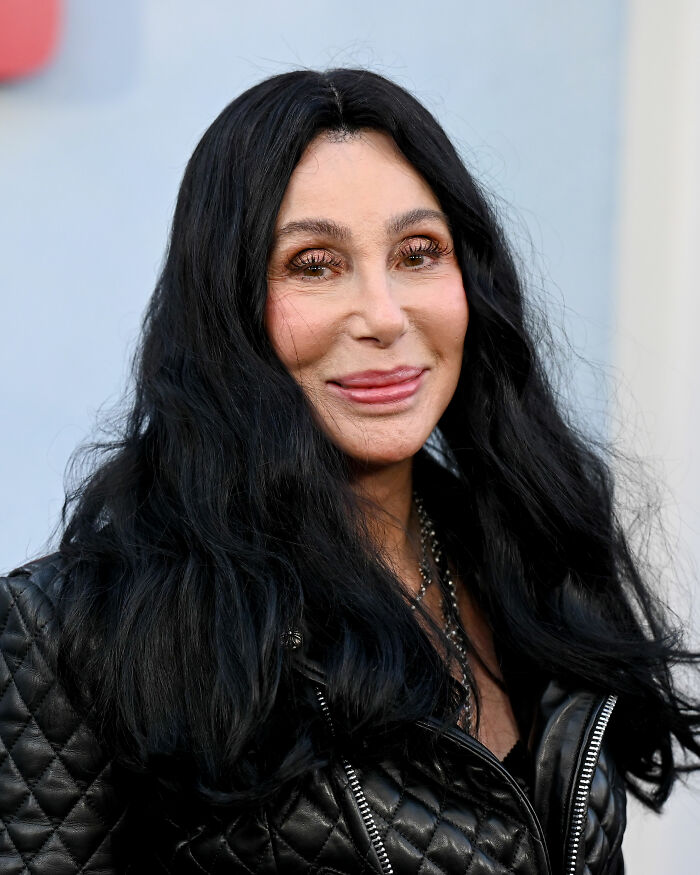 Cher with long black hair and a black quilted jacket, highlighting her melting appearance and plastic surgery concerns.