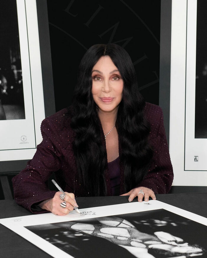 Cher signing a photograph at an event, highlighting concern over her melting appearance and plastic surgery indulgence.