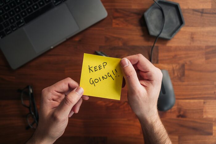 Hands holding a sticky note with keep going message on wooden desk with laptop and mouse, low effort high reward concept.
