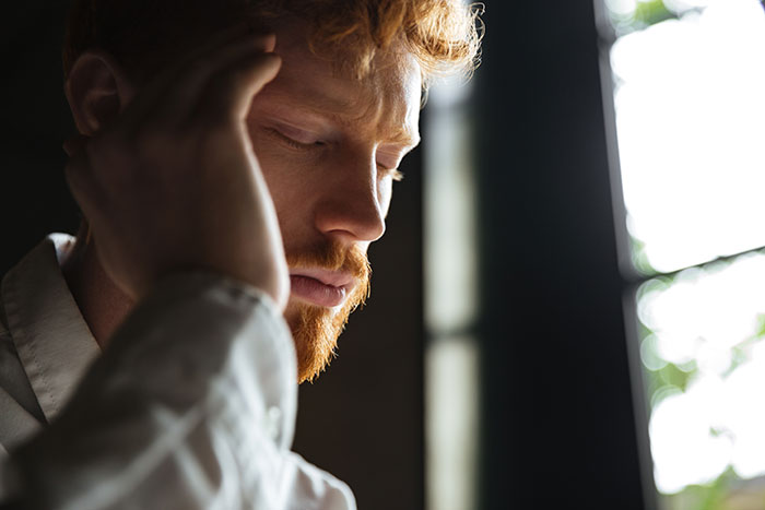 Young man with red hair looking flabbergasted, holding head in hand near window light in a dim room.
