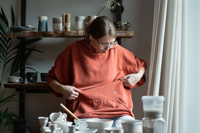 Woman in glasses examining a stain on her sweatshirt, surrounded by broken kitchenware on a cluttered table.