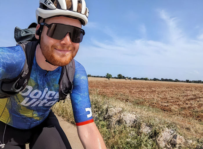 Cyclist wearing helmet and sunglasses riding along rural path with fields under clear blue sky in daytime.