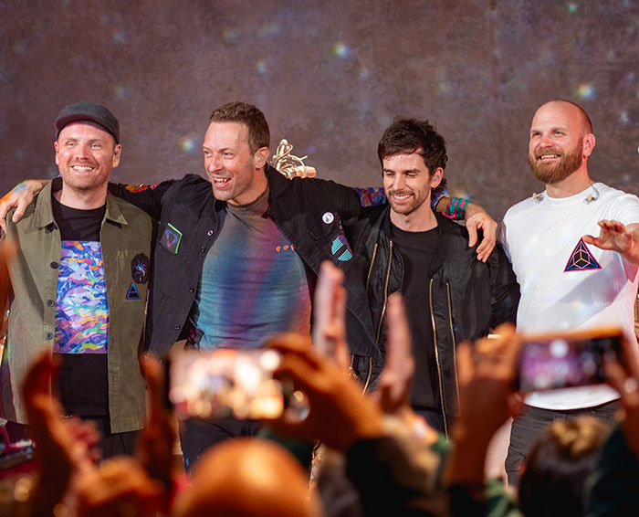 Chris Martin and band members smiling on stage with fans capturing the moment during a live event.