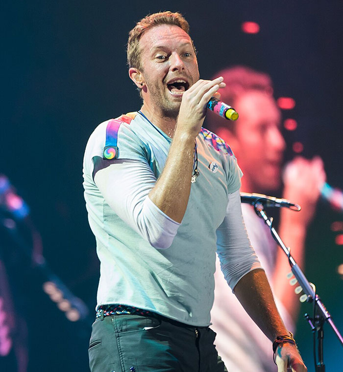 Chris Martin performing on stage, holding a microphone, during a concert amid cheating scandal controversy.