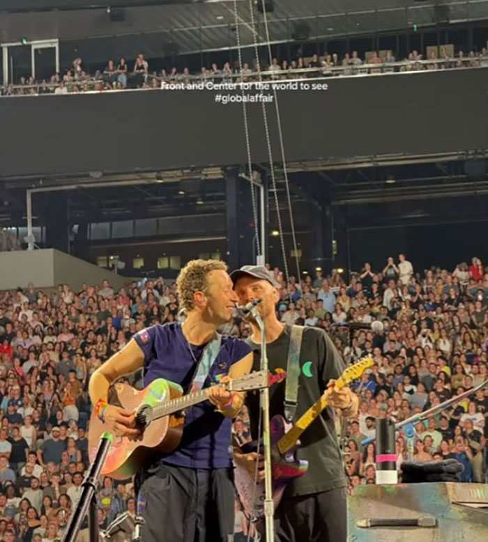 Chris Martin performing live on stage with a guitarist in front of a large concert crowd.
