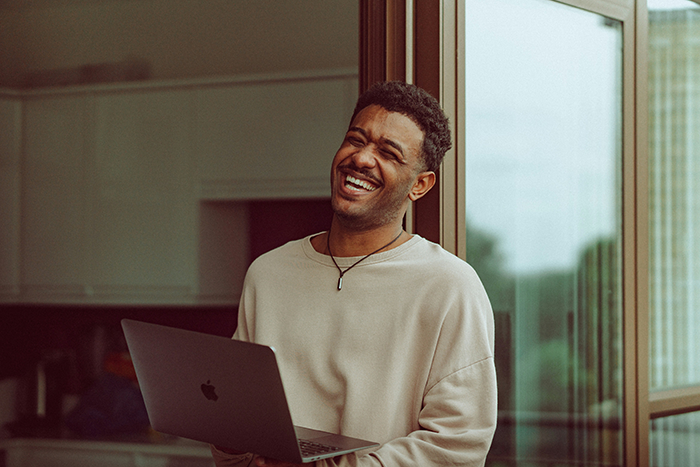 Man holding laptop, smiling and standing by a window, reflecting confidence before giving two weeks notice.