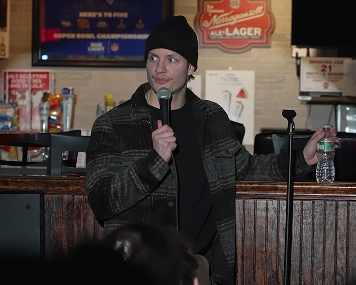 Comedian Matt Rife performing stand-up comedy indoors, wearing a beanie and holding a microphone and water bottle.
