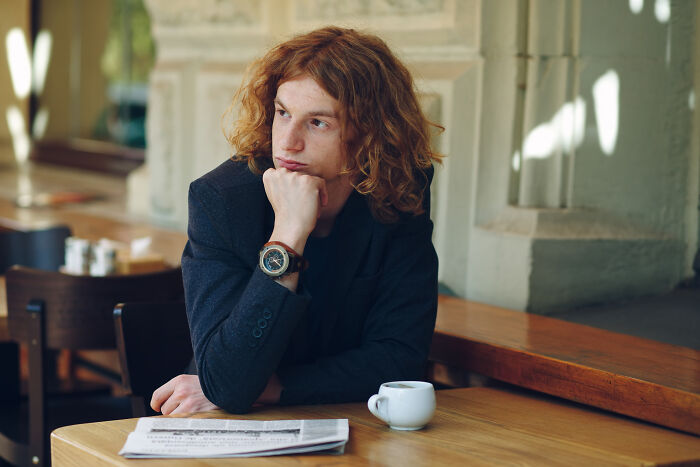 Young man wearing a watch, sitting thoughtfully at a table with a coffee cup and newspaper, concept time facts.