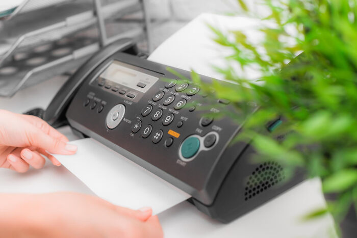 Person sending a fax with a black fax machine on a desk, illustrating concept time facts in office setting.