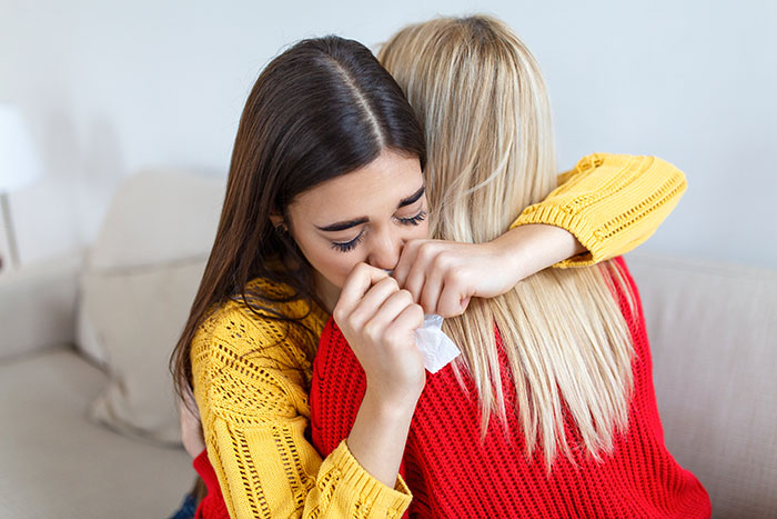 Two women hugging on a couch, one crying and holding a tissue after a racist insult involving black parents at dinner.