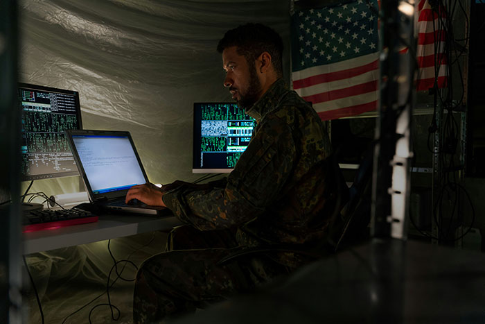 Man in military uniform working on multiple computer screens in a dim room, illustrating conspiracy theorists concept.