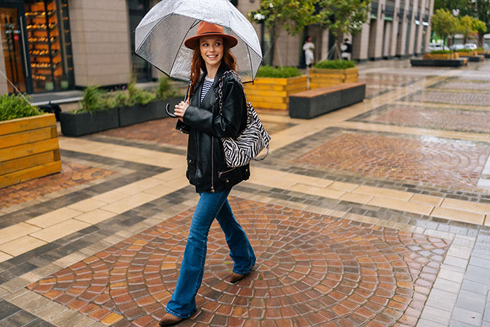 Young woman walking with umbrella on rainy urban street, dressed in hat and leather jacket, illustrating hot take with the boomers concept.