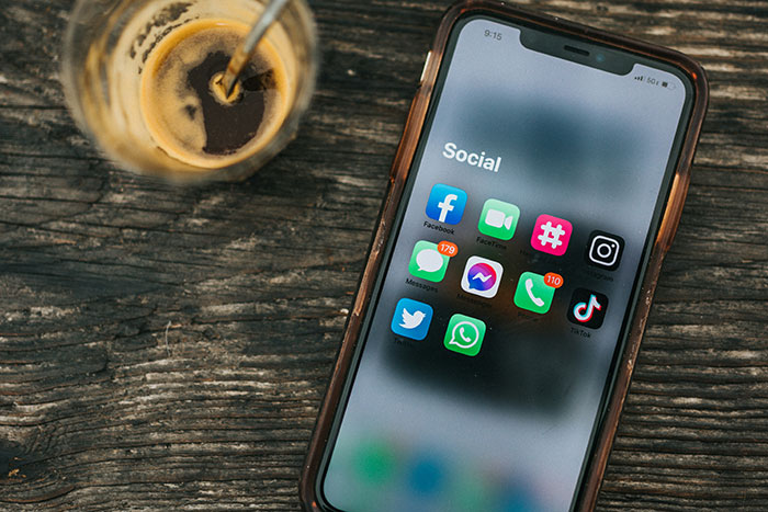 Smartphone displaying social media apps with high notifications, placed on wooden table next to a glass of coffee.