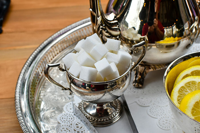Silver sugar bowl filled with sugar cubes on a tray next to lemon slices and a teapot for conspiracy theorists tea setup.