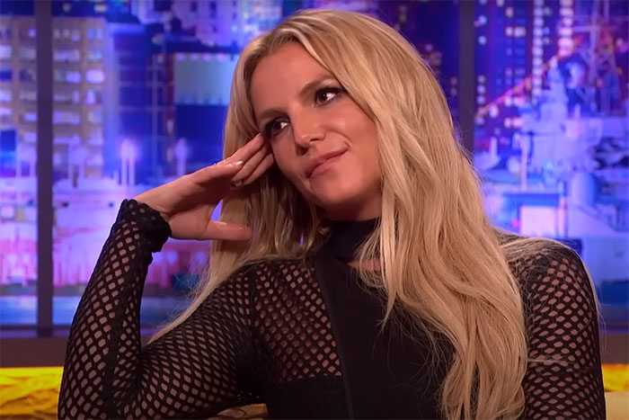 Woman with blonde hair wearing a black mesh top, sitting relaxed in a studio setting, related to conspiracy theorists topic