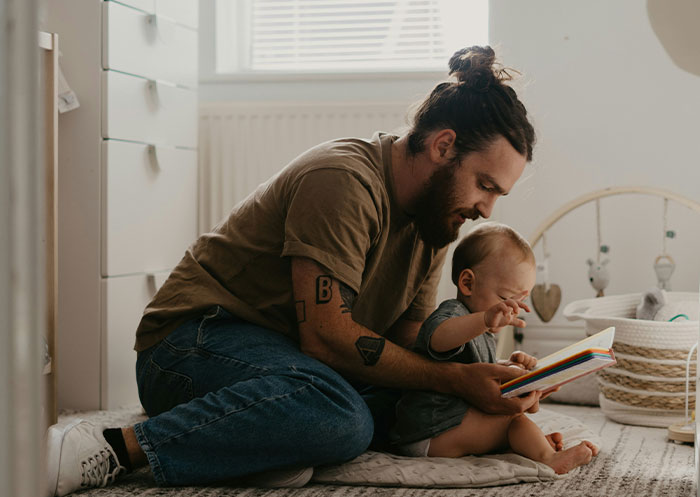 Man with tattoos reading a colorful book to a baby in a cozy room, illustrating ups and downs of a rags to riches partner.
