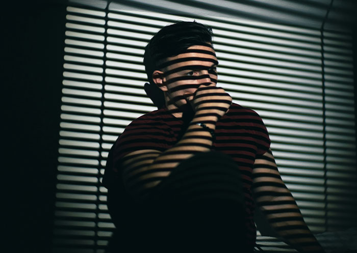Young man sitting in a dark room with striped shadows, reflecting on the ups and downs of a rags to riches partner.