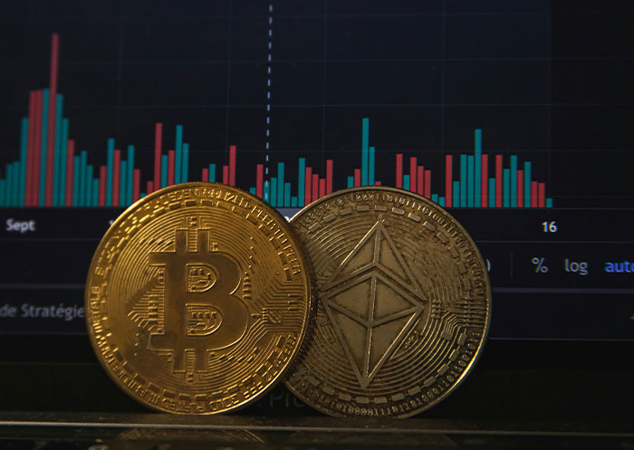 Two golden cryptocurrency coins representing Bitcoin and Ethereum in front of a digital financial chart showing market ups and downs.