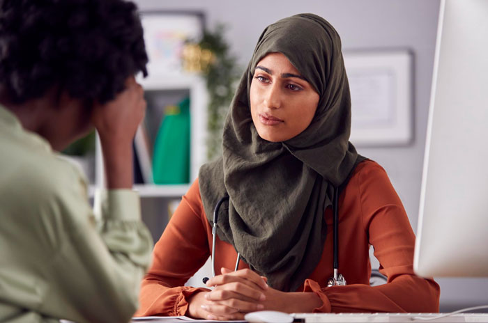 Muslim woman wearing a hijab listening thoughtfully to a coworker in an office, addressing coworker misgenders and religion.