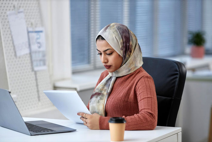 Muslim woman in hijab at office desk looking at documents, addressing coworker misgenders and religion concerns.