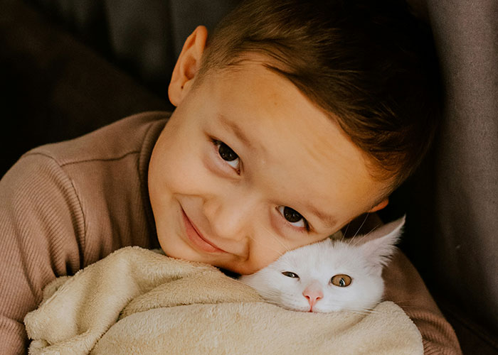 Young boy smiling while hugging a white cat wrapped in a blanket, illustrating lifelong pain from horrible names.
