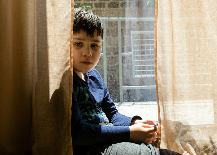 Sad young boy sitting behind curtains at home, illustrating emotional impact of horrible names on children.
