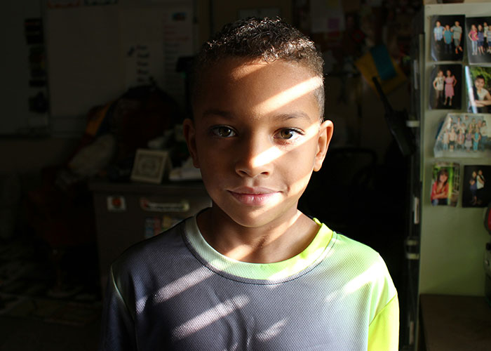 Young boy in a dim room with sunlight stripes on his face, representing children affected by horrible names.