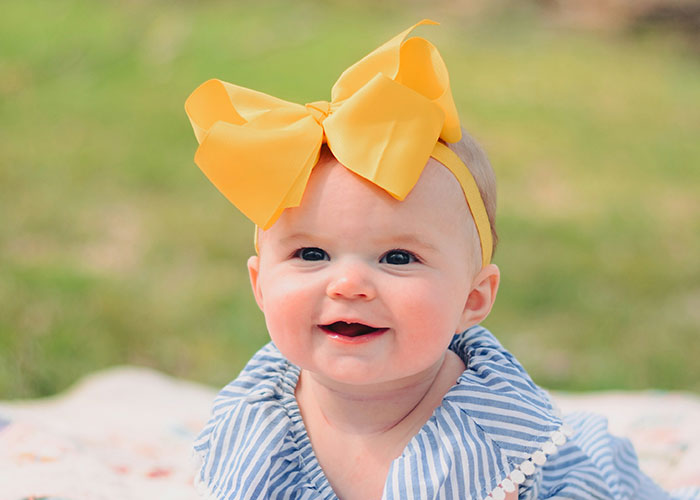 Smiling baby girl wearing a large yellow bow headband outdoors, illustrating examples of horrible names affecting children.