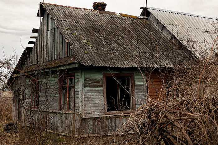 Old abandoned wooden house with broken windows surrounded by dead bushes, evoking creepy vibes in a desolate area.