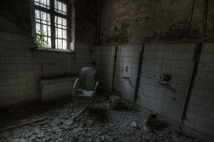 Abandoned room with broken tiles and debris on floor, featuring a rusty chair, giving off creepy vibes.