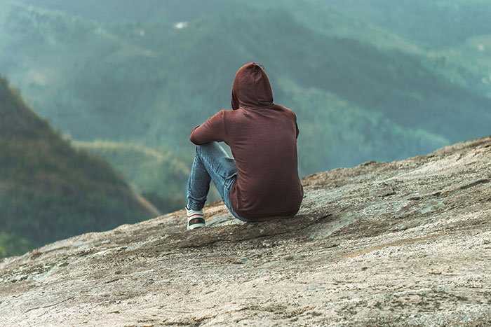 Person wearing a hoodie sitting alone on a rocky ledge outdoors, surrounded by a misty forest landscape.