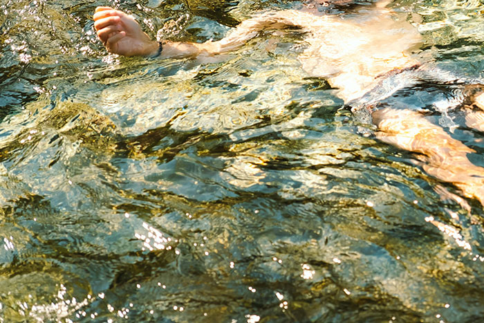 Person's arm floating underwater in a shallow, rocky outdoor stream, evoking creepy and terrifying outdoor scenes.