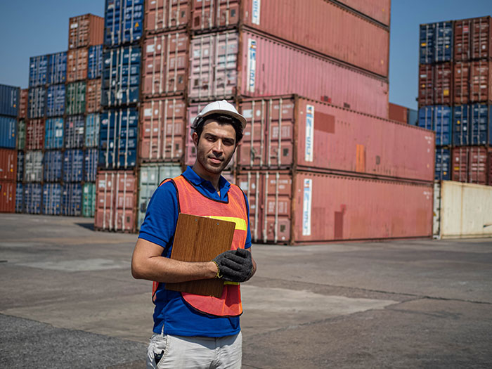 Warehouse worker wearing safety gear and holding a clipboard, standing in front of stacked shipping containers.