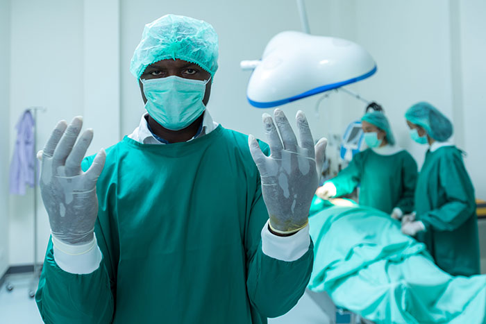 Surgeon wearing surgical scrubs and mask preparing for operation while colleagues attend patient in operating room.