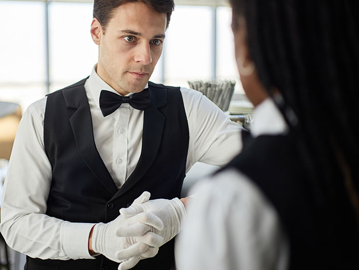Male waiter wearing white gloves and a black vest discusses the creepiest parts of their jobs with a colleague.